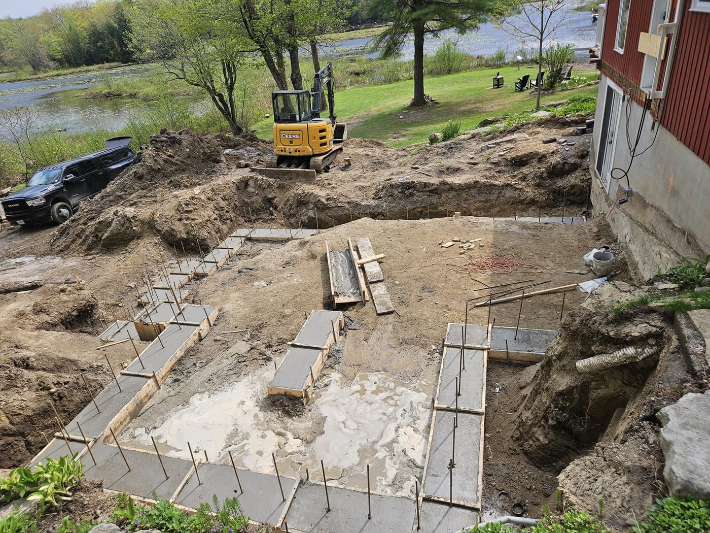 Concrete footings formed in level runs and steps down a sloped lot beside an existing house, with rebar dowels standing up from the forms and water beyond the lawn
